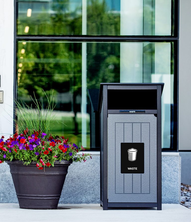 Outdoor waste bin with modern gray panel design placed beside a flower planter in front of a glass building.