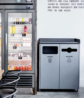 indoor dual-stream waste and recycle bin beside a refrigerated beverage display in a modern café.