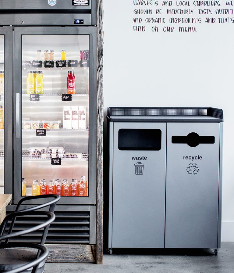 indoor dual-stream waste and recycle bin beside a refrigerated beverage display in a modern café.