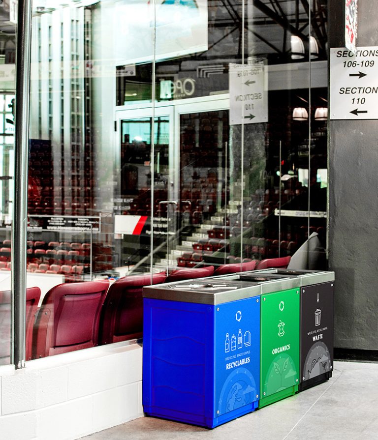 Indoor recycling and waste station with blue, green, and black bins for recyclables, organics, and waste placed near arena seating