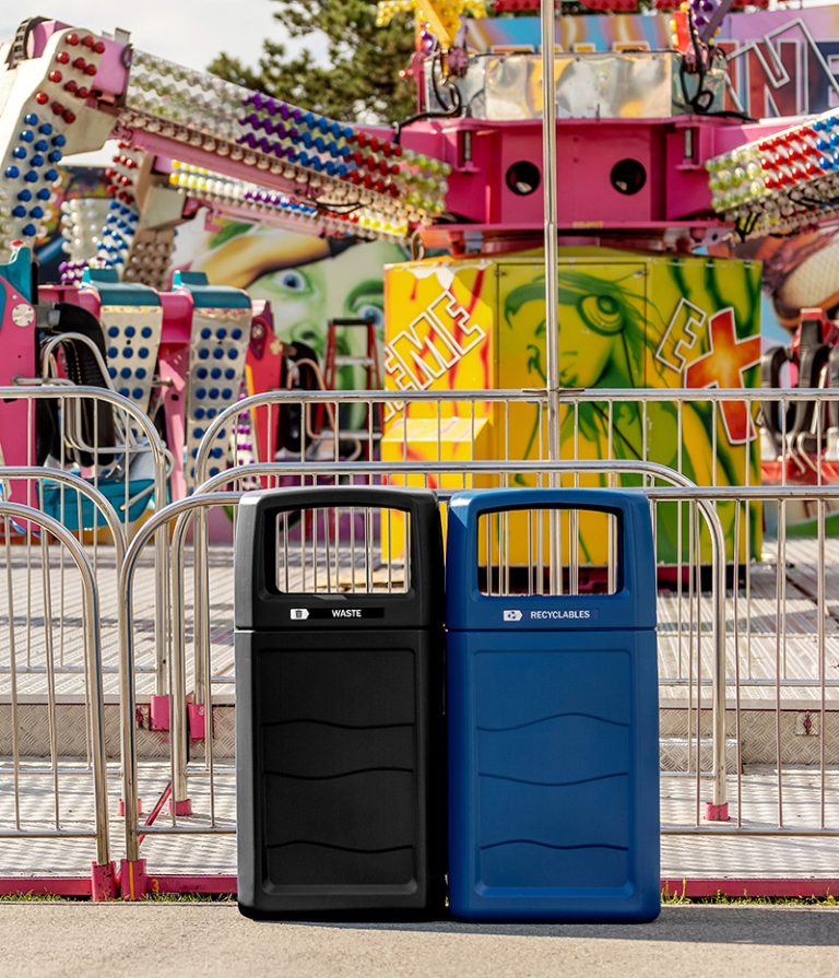 Outdoor waste and recycling bins positioned near a colorful amusement park ride with bright lights and railings.