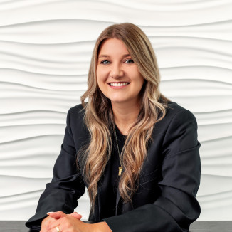 Smiling woman with long, wavy hair in a black blazer sits at a table with a wavy white background.