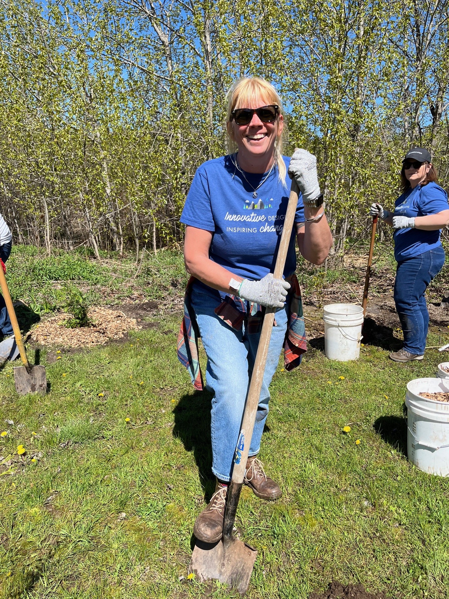 Smiling woman with sunglasses holds a shovel while gardening outdoors with others on a sunny day.