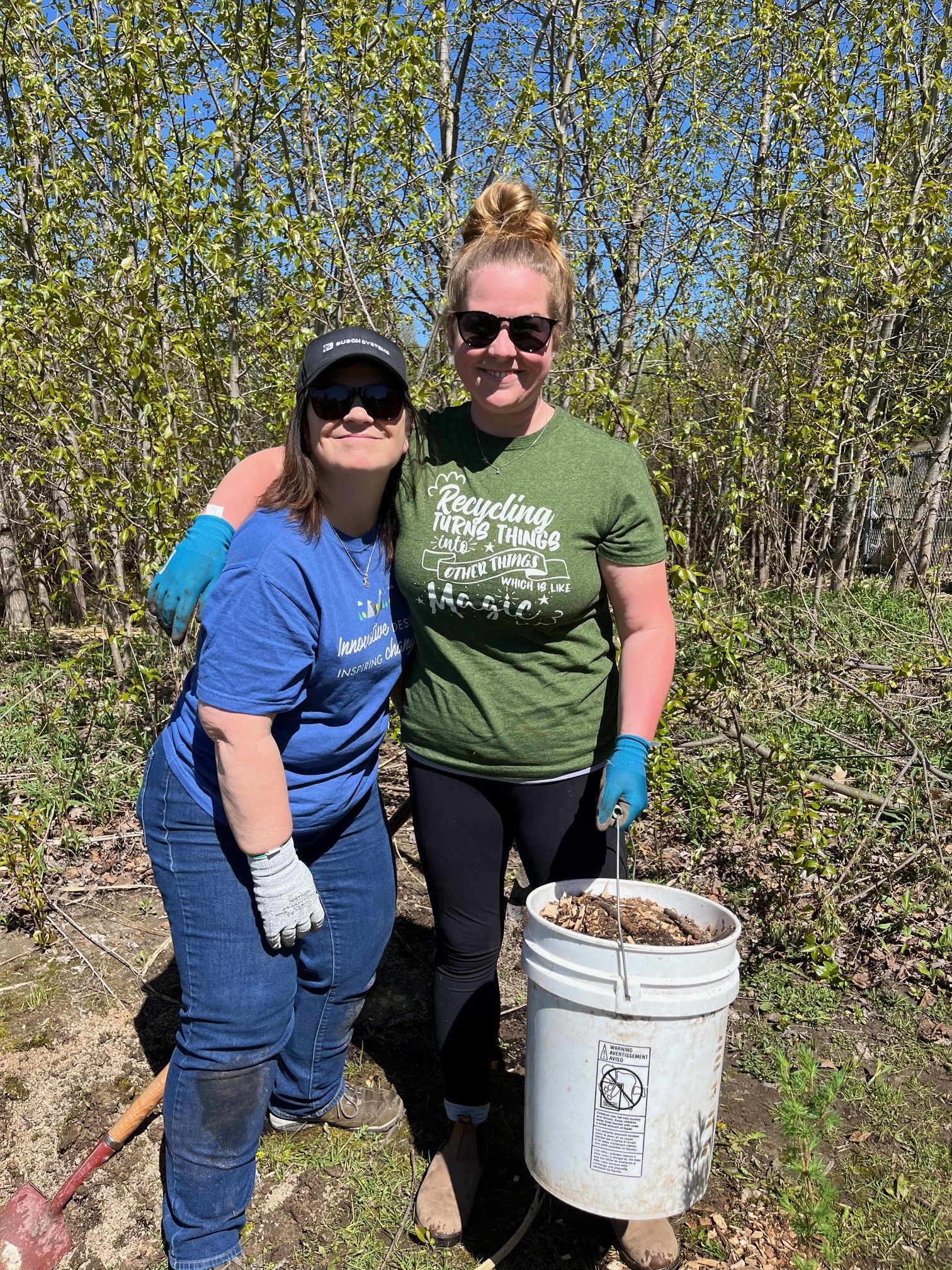 Two women smiling outdoors, wearing gloves and holding a bucket of leaves while gardening.