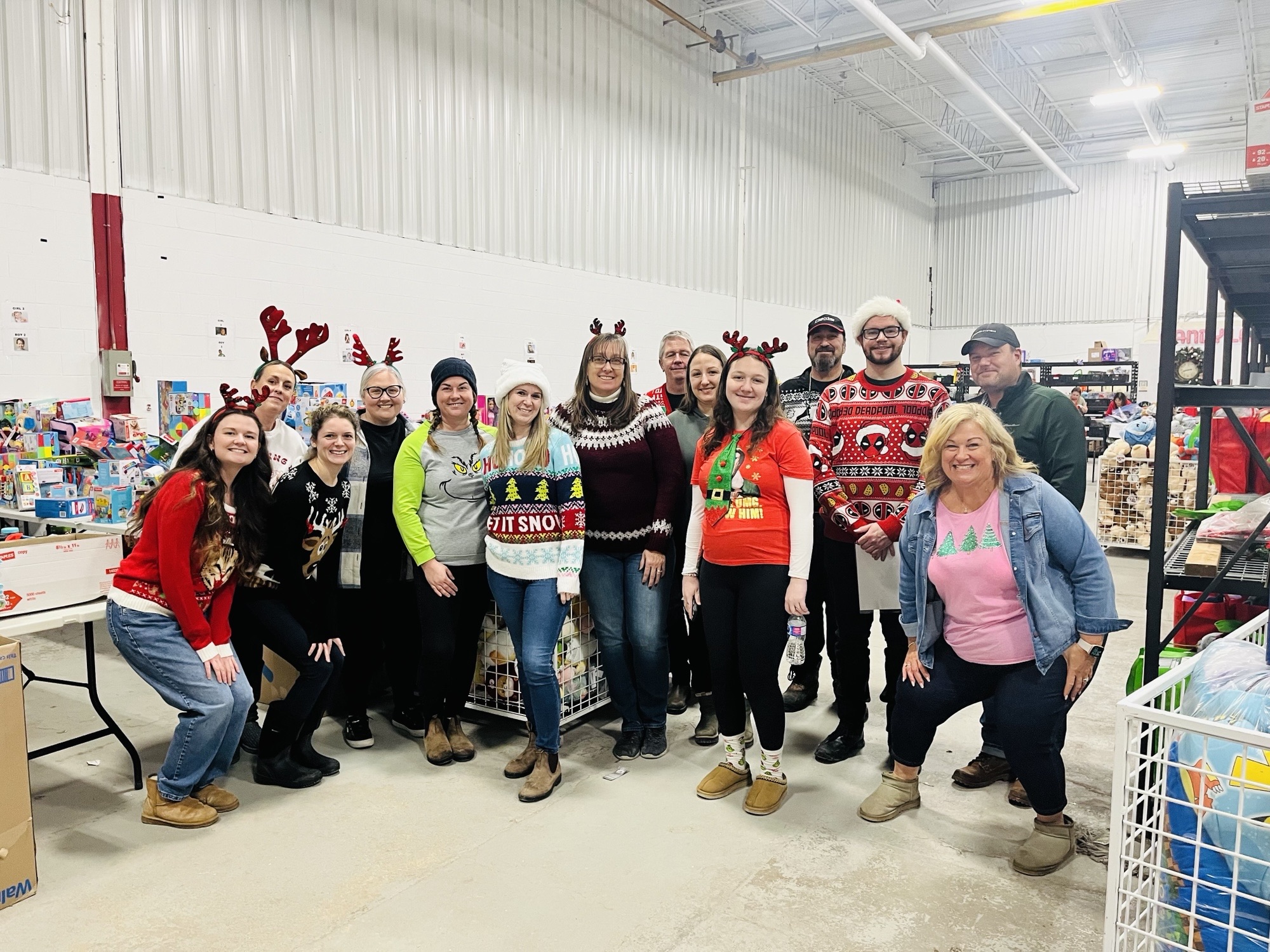 Group of people wearing festive holiday sweaters and accessories posing together during a community event inside a warehouse.