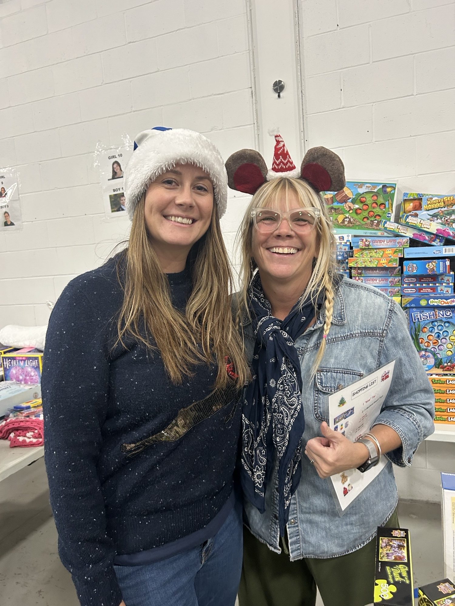 Two people wearing festive holiday hats and smiling during a community gift event surrounded by colorful toys.