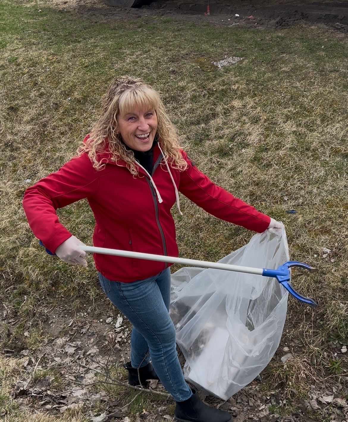 Woman in a red jacket picks up litter with a grabber tool and holds a trash bag outdoors on grassy ground.
