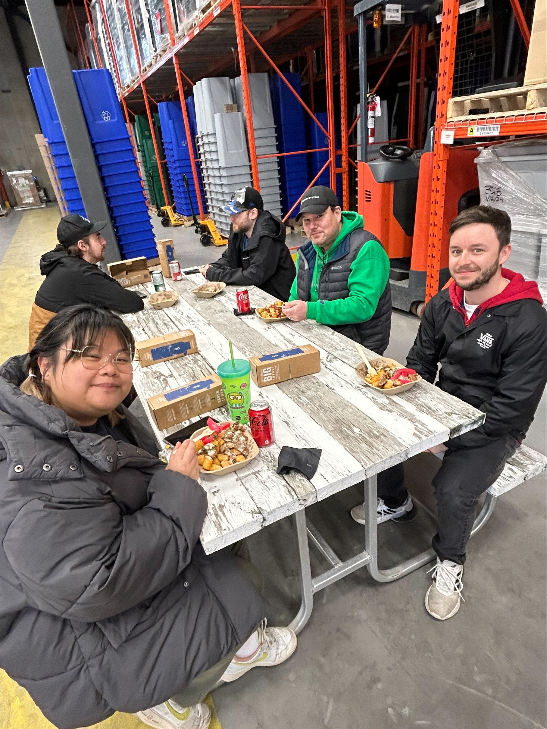 People sitting together at a wooden table enjoying lunch inside a warehouse setting with recycling bins in the background.