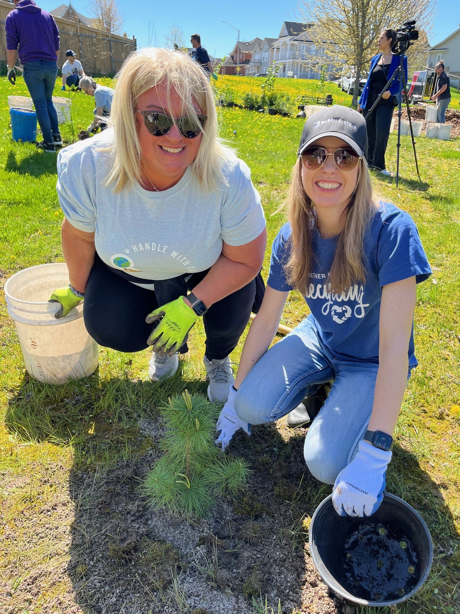 Two women kneel on grass, holding buckets and smiling while planting a small tree on a sunny day.