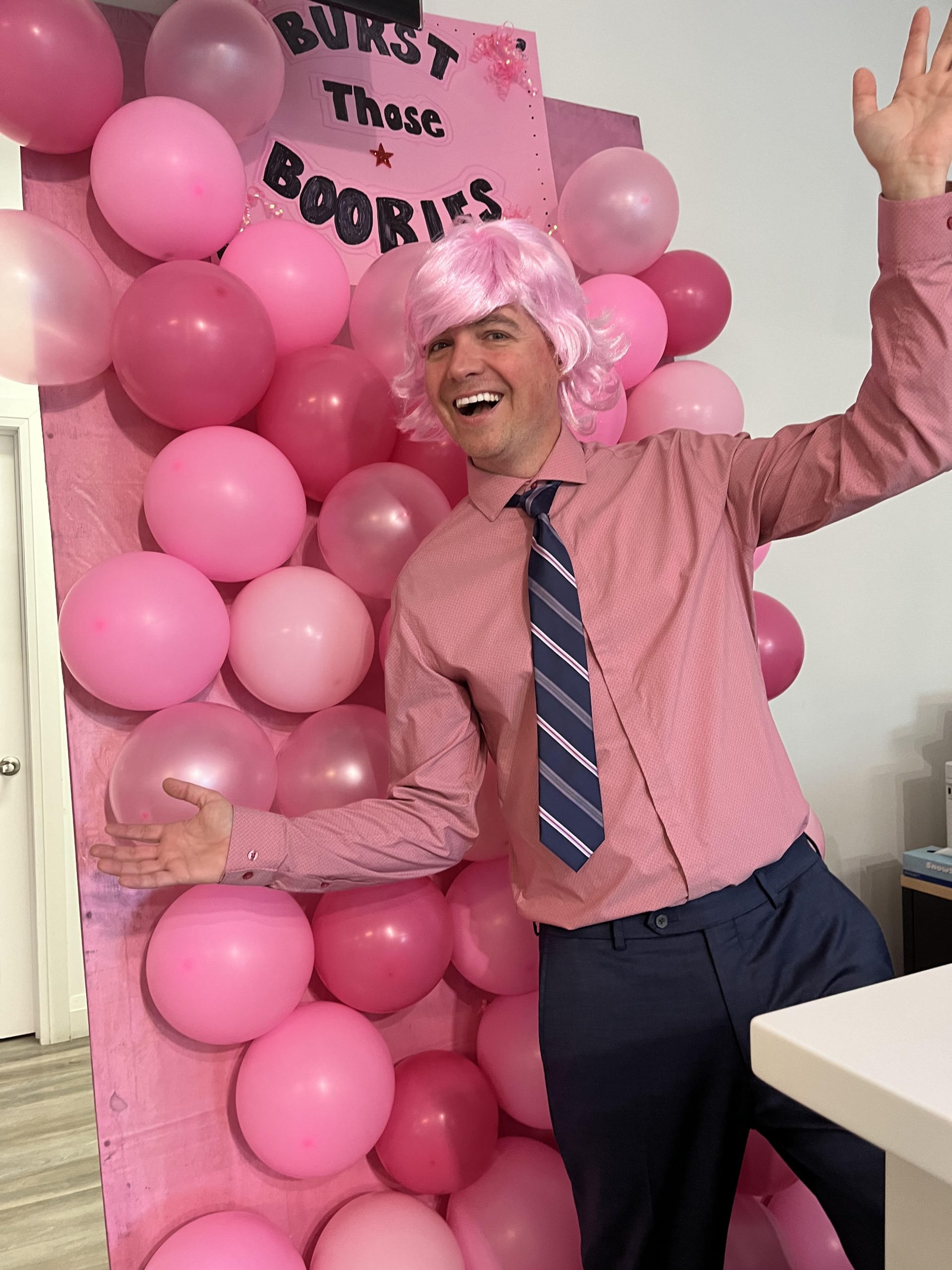 Person wearing a pink wig and shirt posing playfully in front of a wall decorated with pink balloons for a themed event.