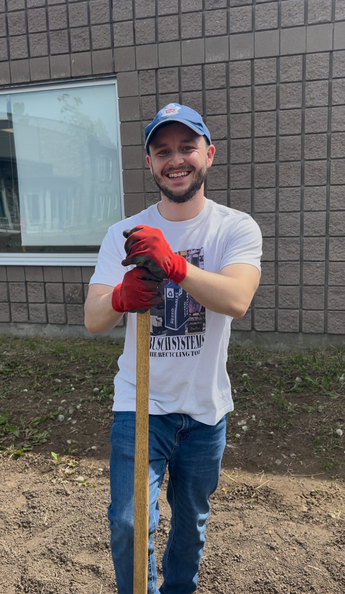 Smiling man in red gloves holds a wooden tool, standing outside near a building with a large window.