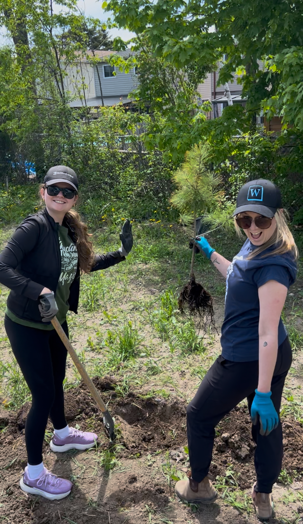 Two women planting a tree outdoors, smiling at the camera and holding gardening tools and a sapling.