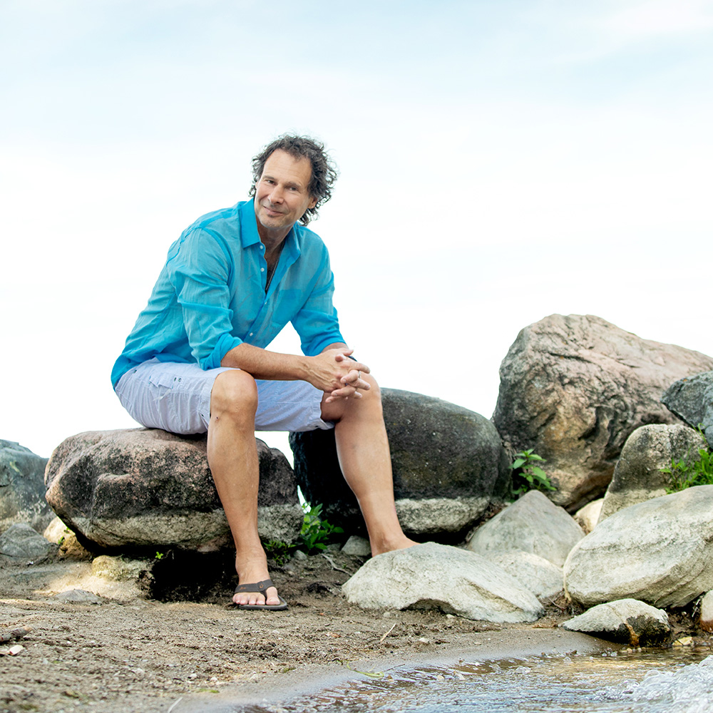 Craig Bush sitting on large rocks outdoors near the water, wearing a turquoise shirt and white shorts, looking relaxed in a natural setting