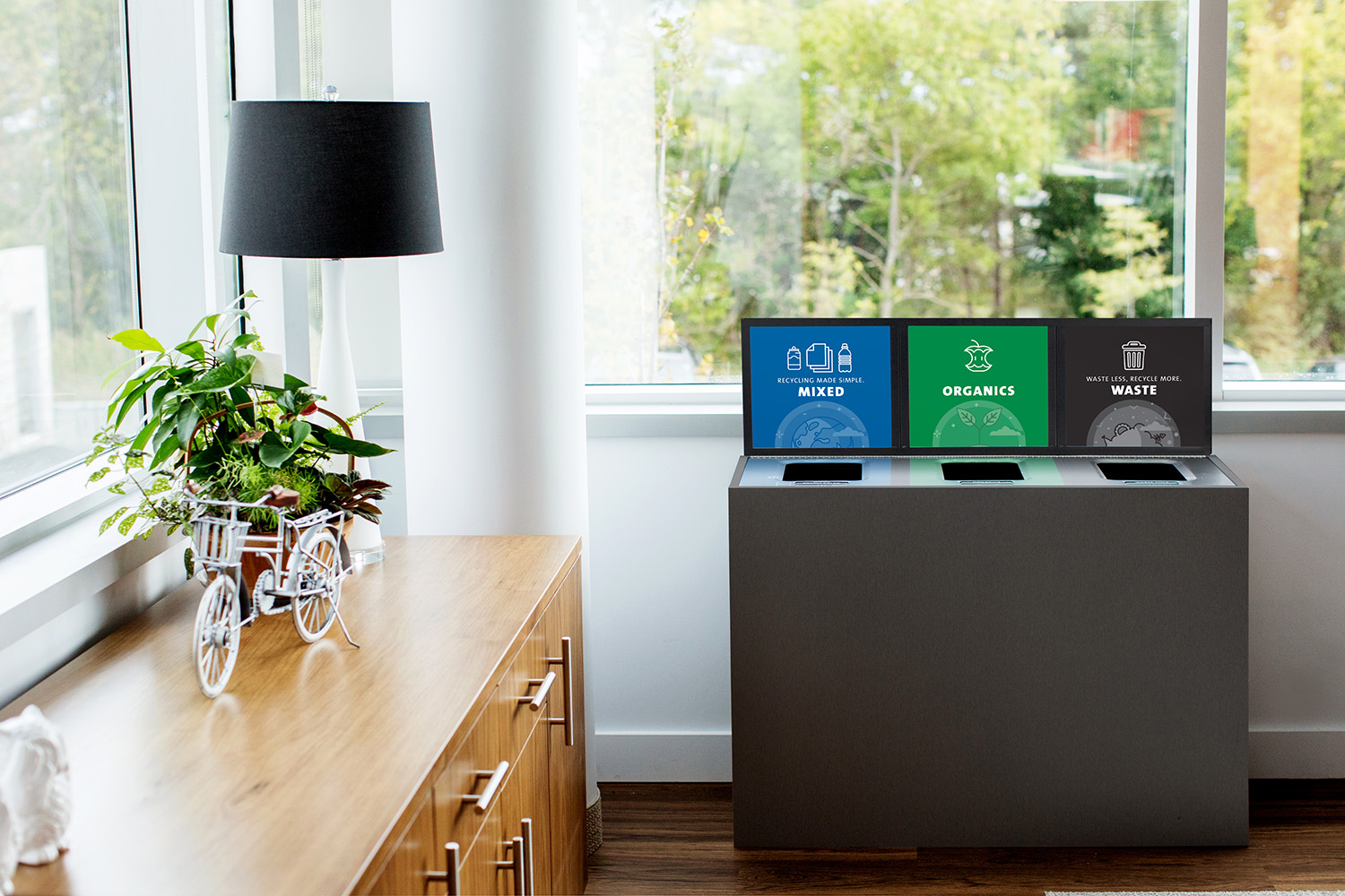 A modern indoor recycling station with separate bins for mixed, organics, and waste stands next to a window, beside a wooden cabinet holding a decorative plant in a tricycle-shaped planter.