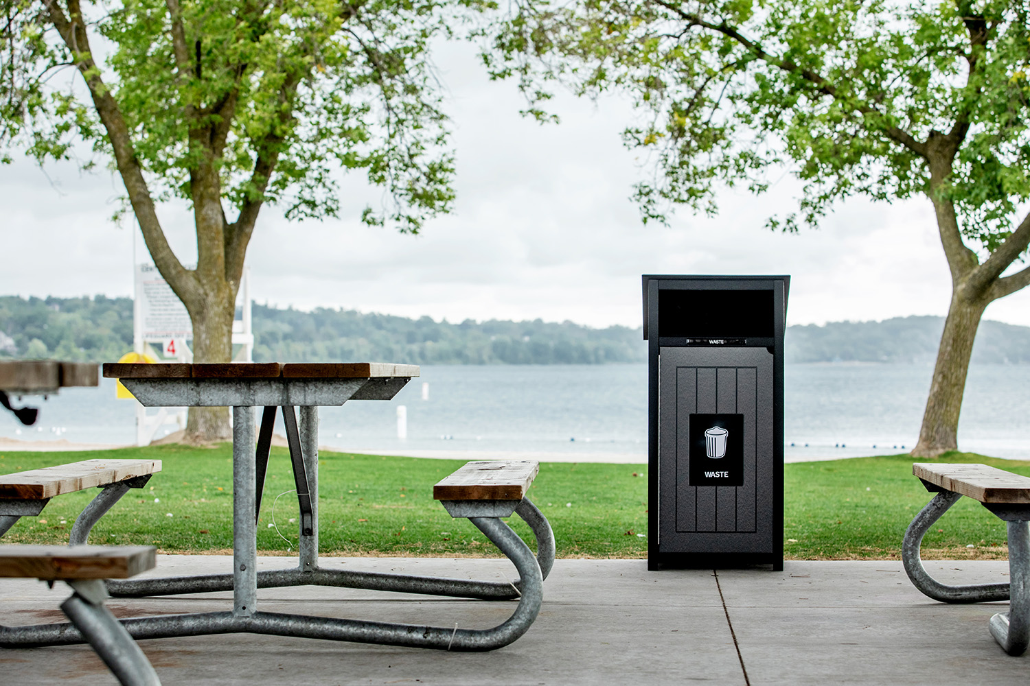A black waste bin stands on a concrete path between empty picnic tables near a lake, with green grass, trees, and a cloudy sky in the background.