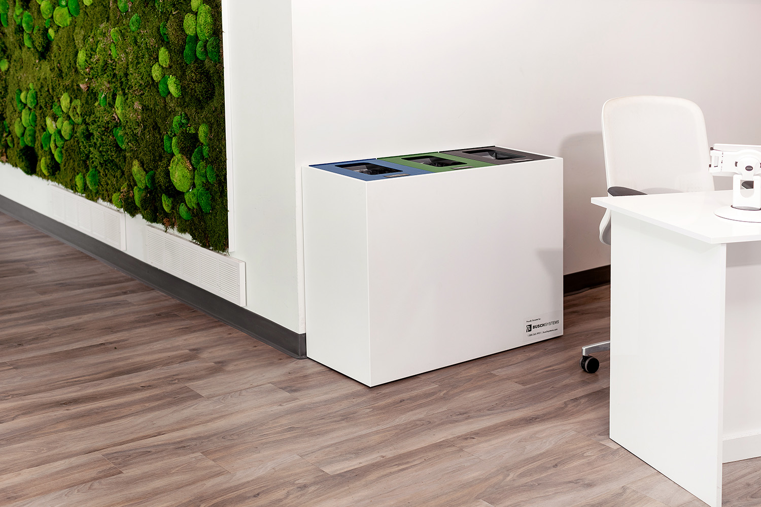 A modern office space with a white three-bin recycling station next to a white desk and chair; a wall with green moss decor is visible in the background.