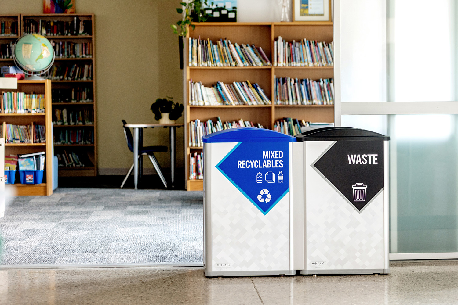 Two bins labeled Mixed Recyclables and Waste stand side by side in a library, with bookshelves and a globe visible in the background.