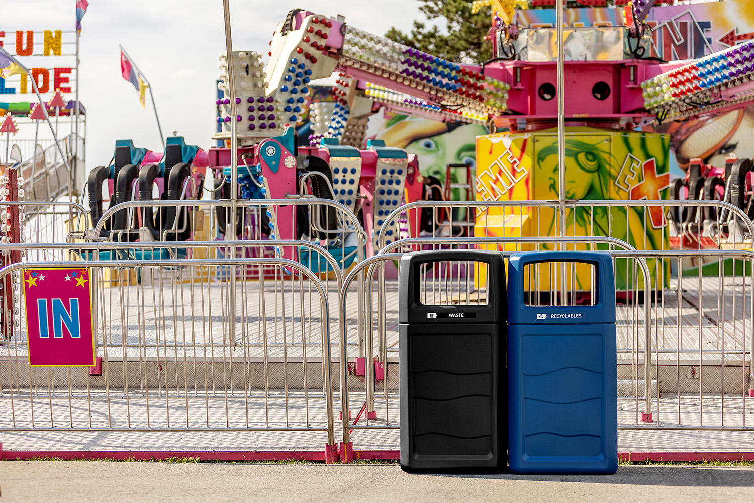 Two trash bins in front of a colorful, empty amusement park ride, with metal barriers in the foreground.