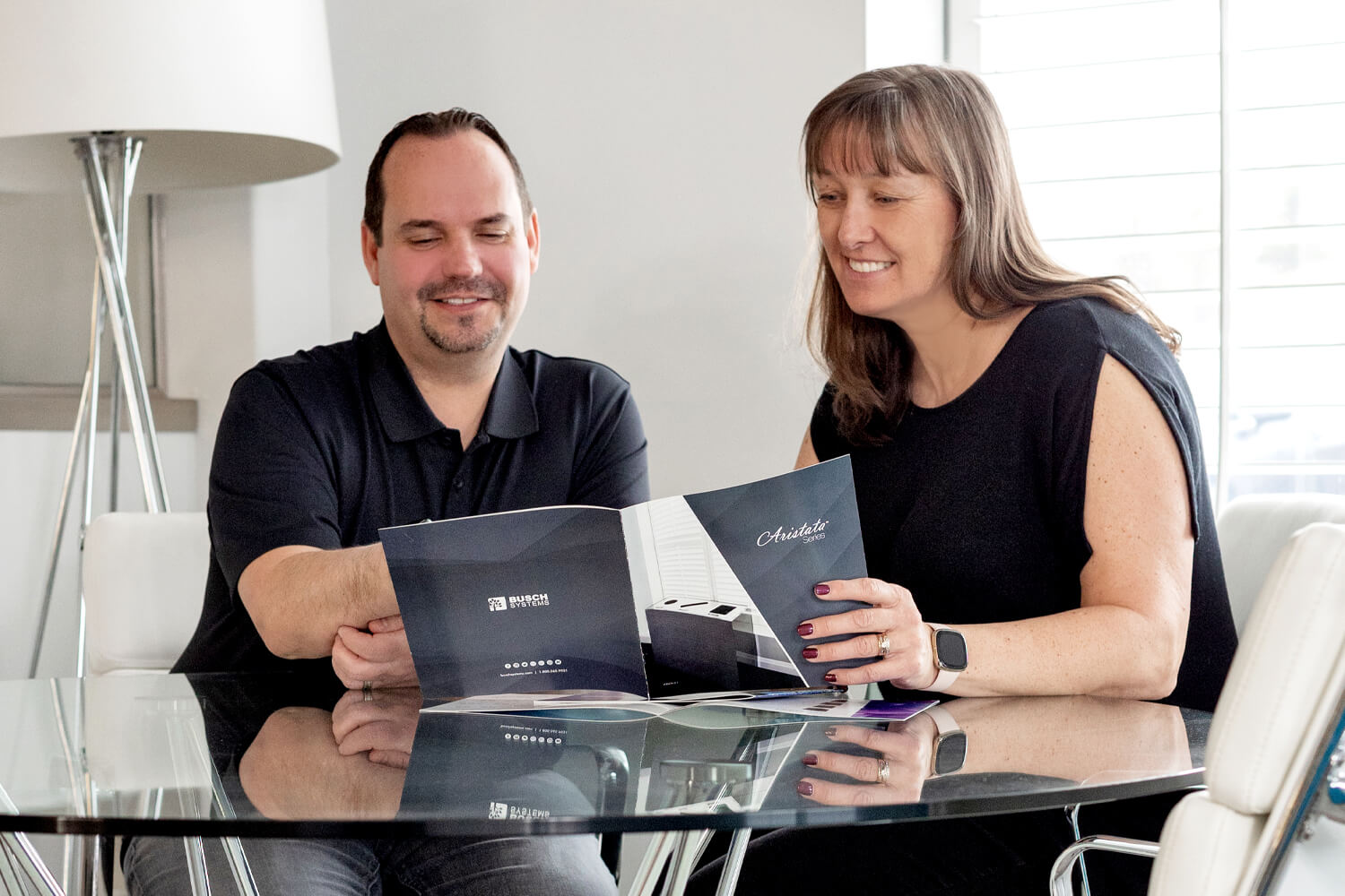 A man and woman sit at a glass table, smiling while looking at a brochure together in a bright, modern room with a lamp and white chairs.