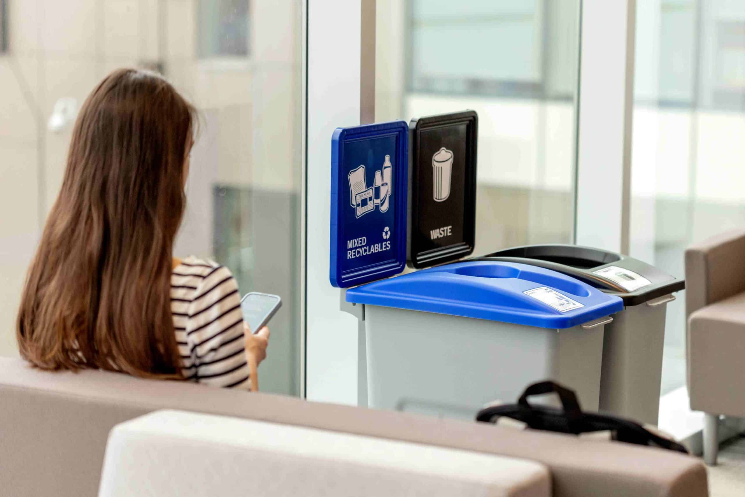 woman on her phone sitting beside recycling and waste containers